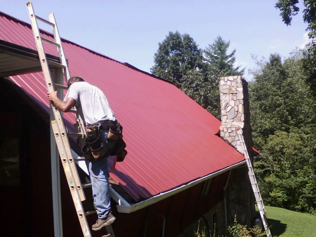 Roofer on a ladder beside a red metal roof and stone chimney — Soffit & Fascia Repair by Roofing Brothers in Carrboro